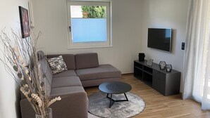 A modern living room with a sectional sofa, a small round table, a flat-screen TV, and a window with blinds. - Modern apartment with large balcony. Near city center. (Meran)