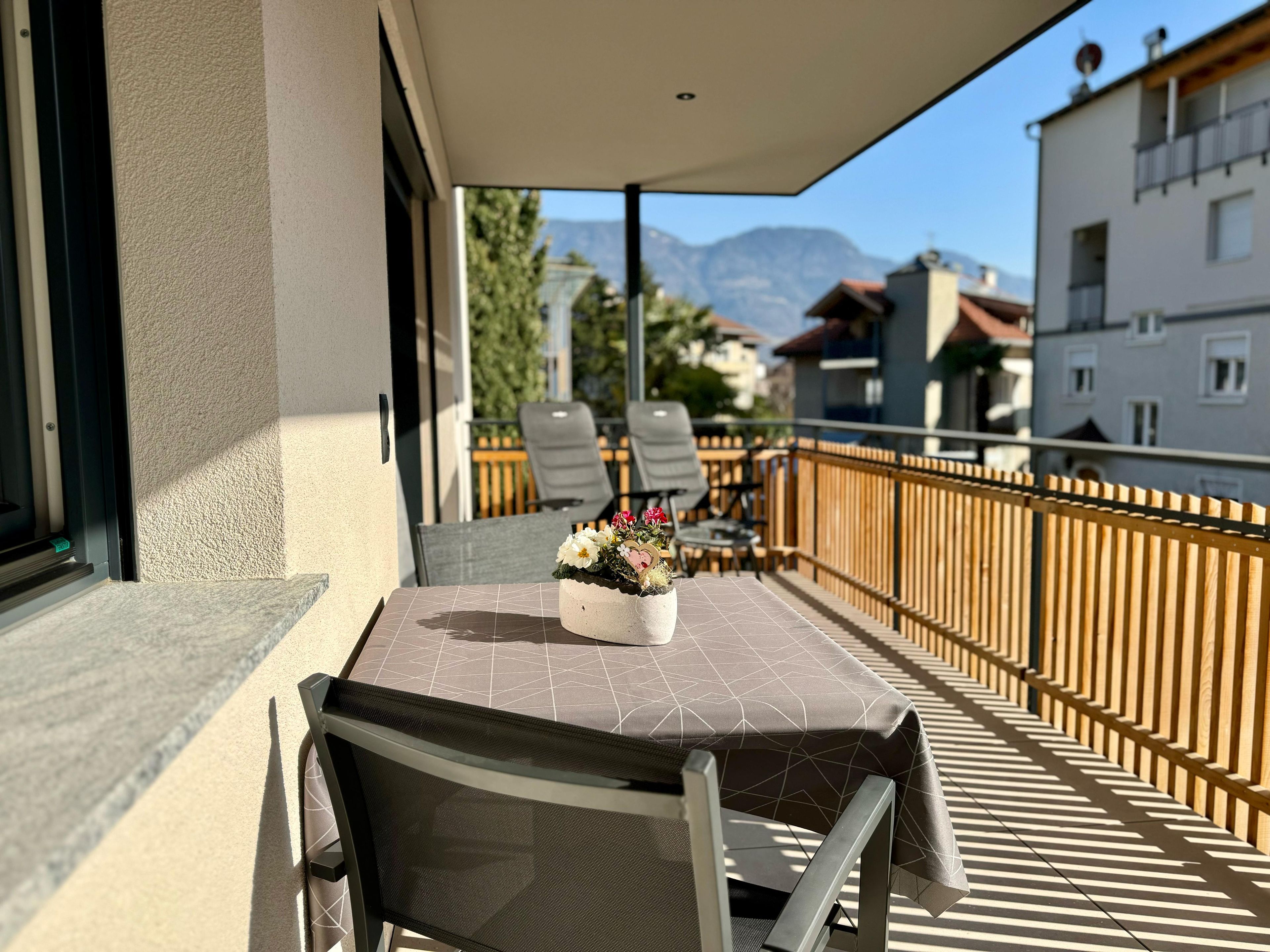 Un balcón con mesa y sillas, un florero y vistas a edificios y montañas.