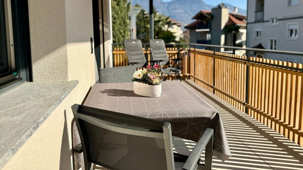 A balcony with a table and chairs, a flower vase, and a view of buildings and mountains. - Modern apartment with large balcony. Near city center. (Meran)