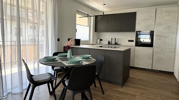 A modern kitchen with a central island, dark cabinetry, and a dining area with a table set for four. - Modern apartment with large balcony. Near city center. (Meran)