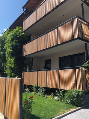 Un immeuble à plusieurs étages, doté d’un balcon à chaque niveau, entouré de verdure et d’un jardin.