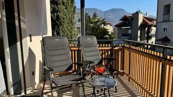 Ein Balkon mit zwei Stühlen und einem Tisch, einem Holzzaun und Blick auf Gebäude und Berge im Hintergrund.