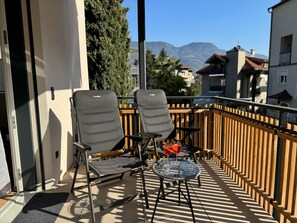 Un balcon avec deux chaises et une table, une clôture en bois et une vue sur des bâtiments et des montagnes en arrière-plan.