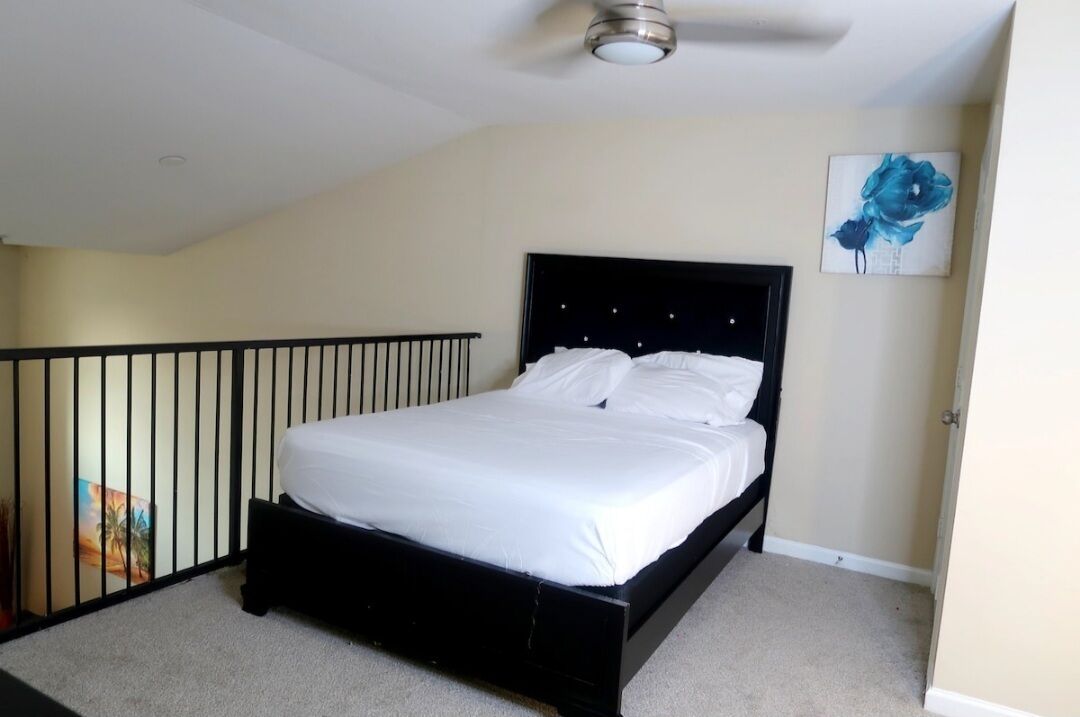 Une chambre avec un cadre de lit noir, du linge de lit blanc, un ventilateur de plafond et un tableau représentant une fleur bleue accroché au mur.