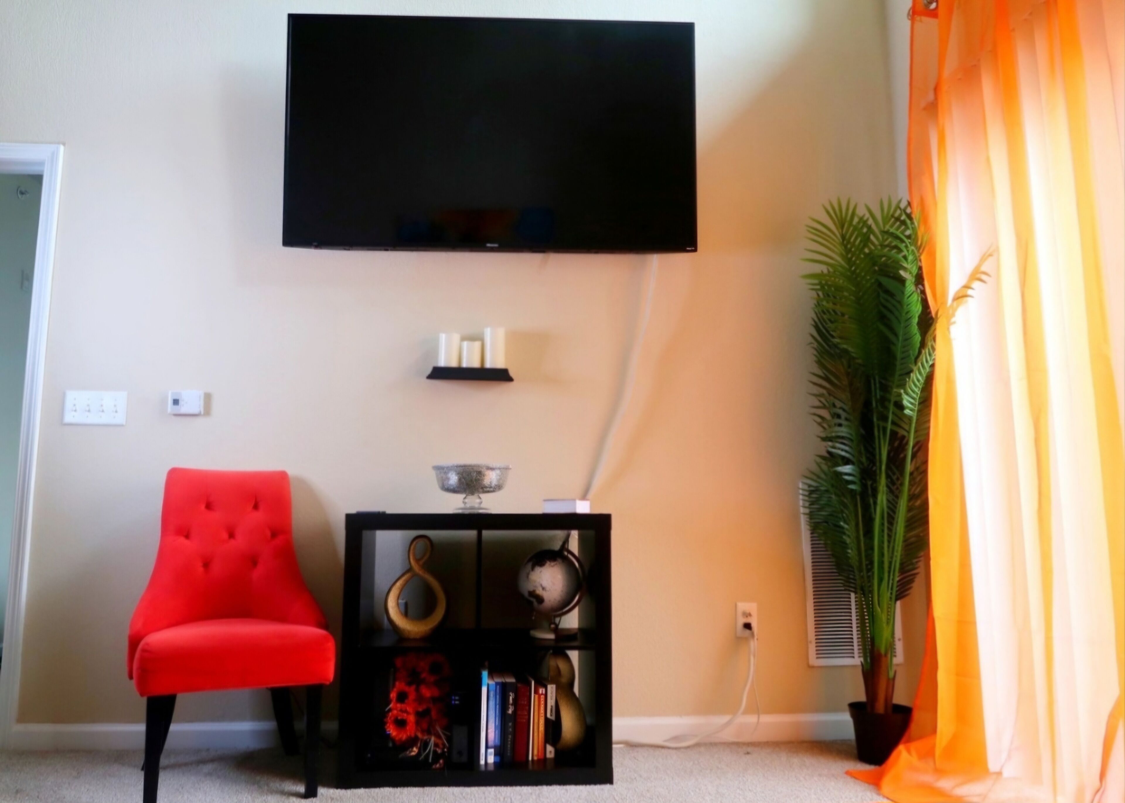Una sala de estar con un televisor de pantalla plana montado en la pared, un sillón rojo, una mesita de noche negra con objetos decorativos y una planta en maceta.
