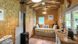 En mysig stuga med en vedeldad kamin, en säng med kuddar, ett fönster och en liten köksdel.