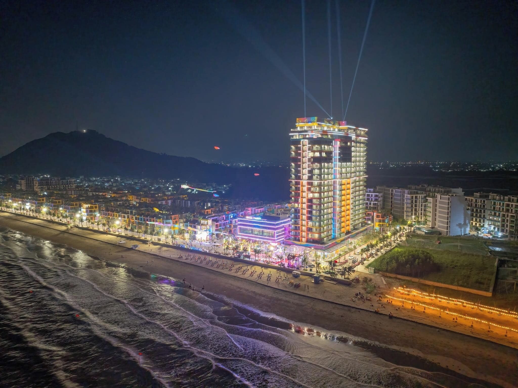 A brightly lit hotel with multiple floors and a prominent central tower, situated by a beach with a water body in the foreground.