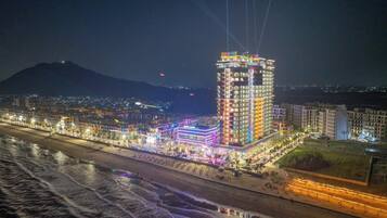 A brightly lit hotel with multiple floors and a prominent central tower, situated by a beach with a water body in the foreground.