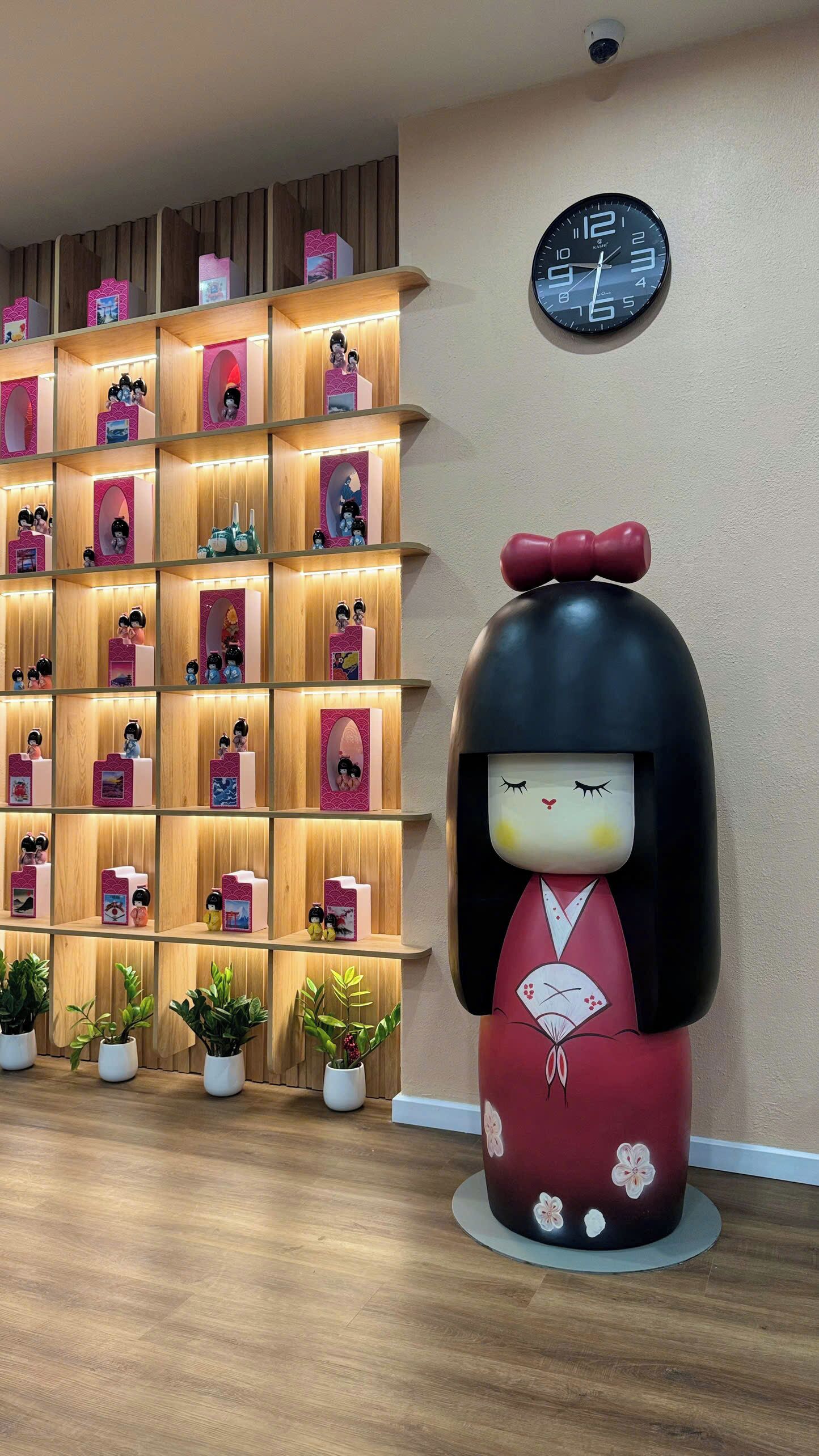 A room with a wooden shelf displaying figurines, a large decorative doll, a clock, and potted plants.