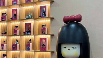 A room with a wooden shelf displaying figurines, a large decorative doll, a clock, and potted plants.
