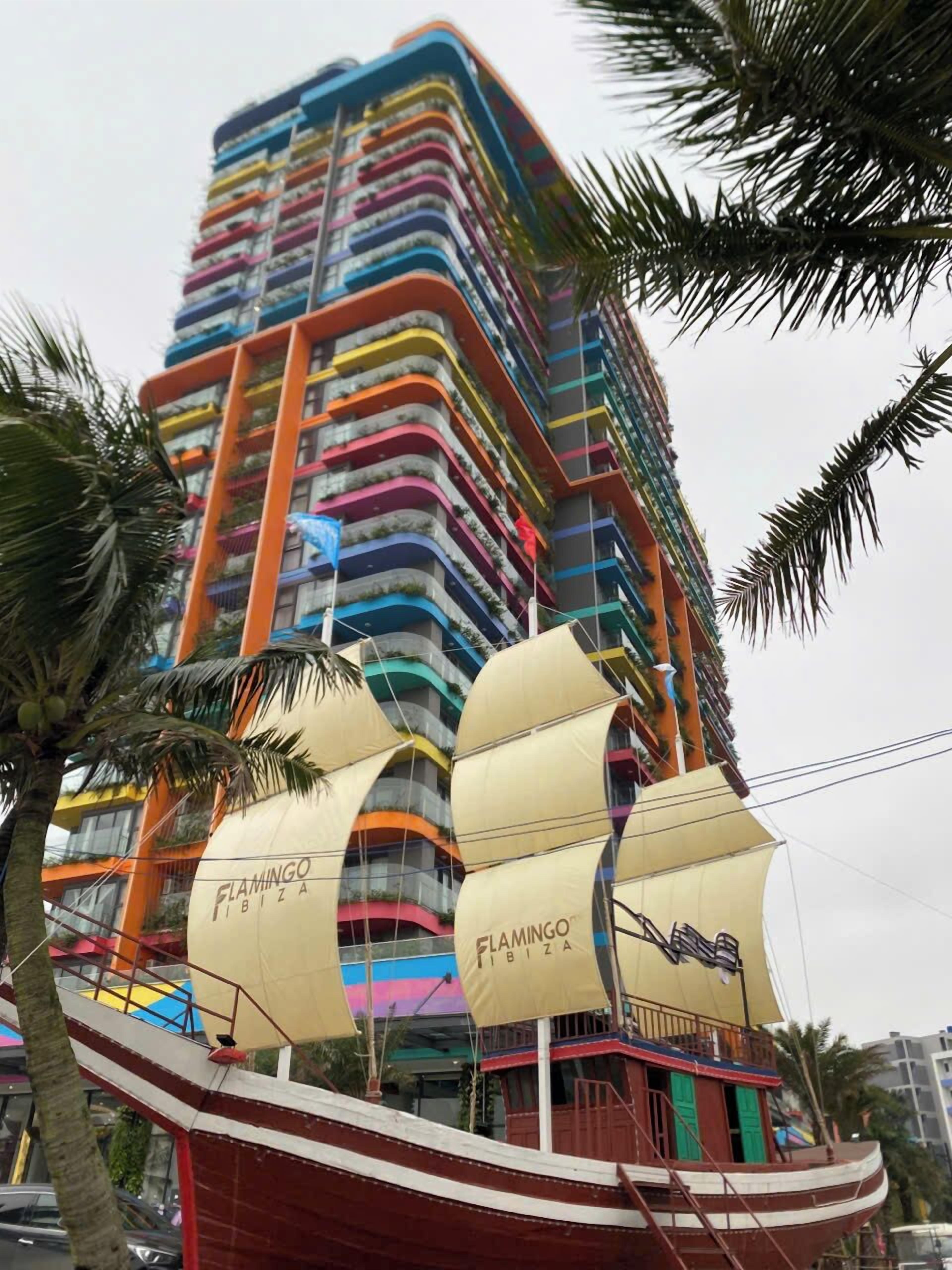 Un immeuble à plusieurs étages doté de balcons, une structure en forme de bateau à l’avant et des palmiers.