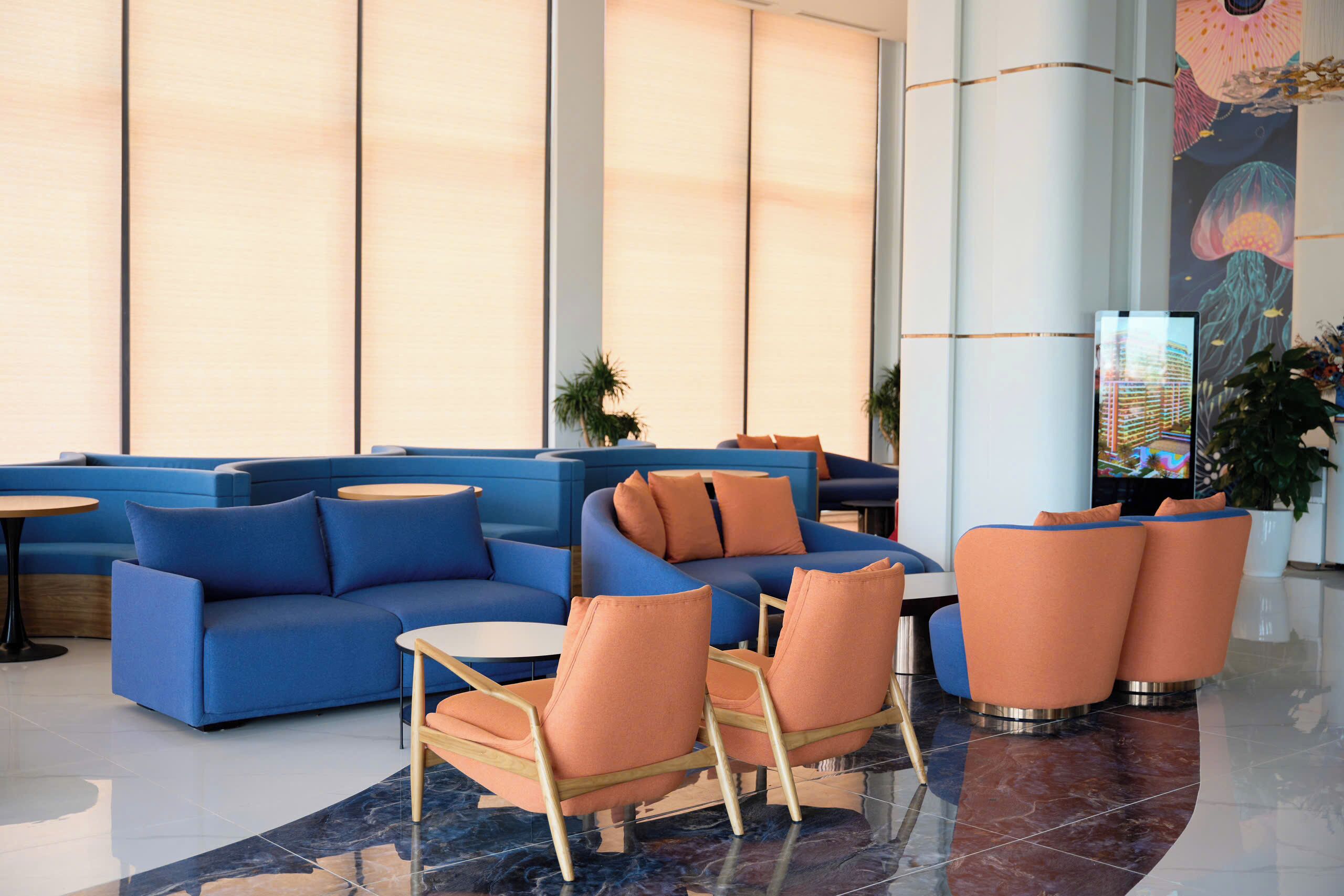 Un vestíbulo moderno con sofás azules, sillones naranjas y una amplia pantalla digital.