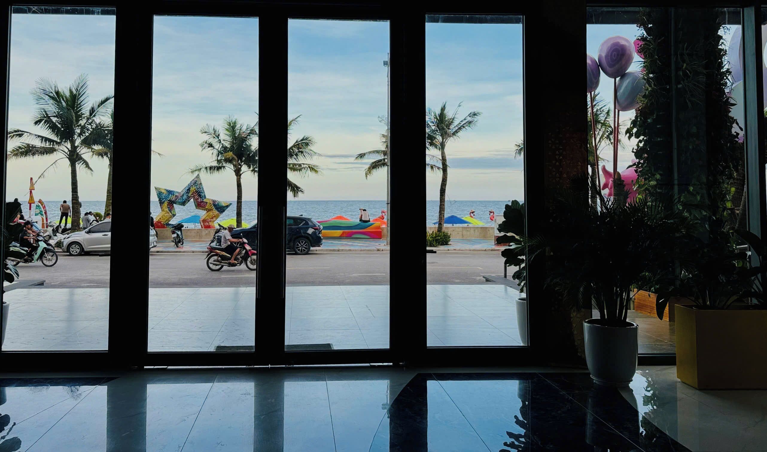 A view from inside a building with large windows overlooking a beach. There are palm trees, a colorful star-shaped decoration, and a few people and vehicles on the street.