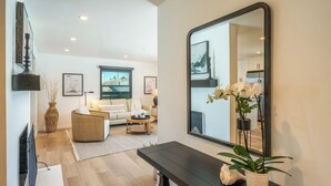 Un salon moderne avec un canapé, une table basse et un grand miroir. Il y a un téléviseur à gauche et une plante en pot à droite.