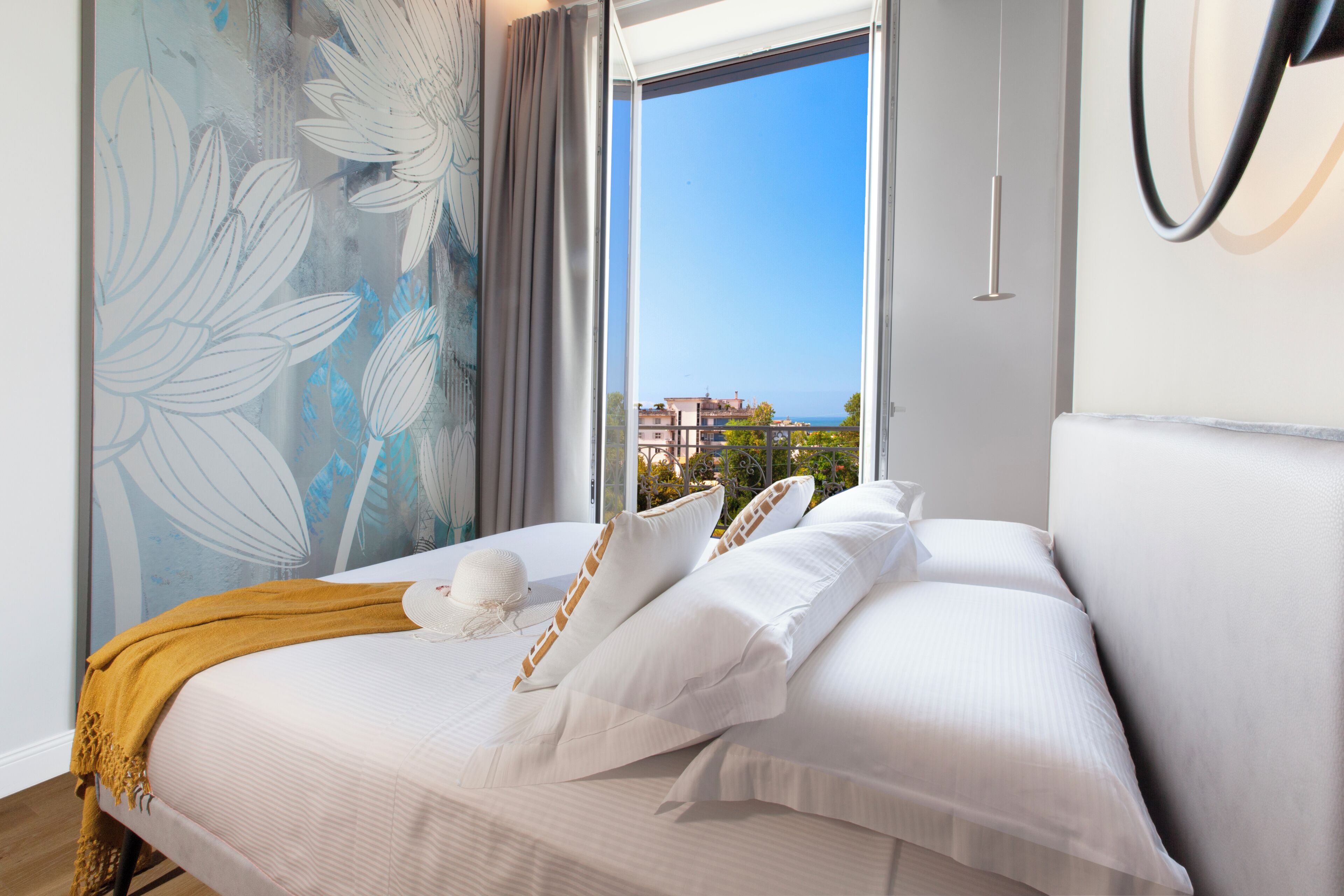 Habitación de hotel con una cama grande, ropa de cama blanca, vista al mar y un mural en la pared.