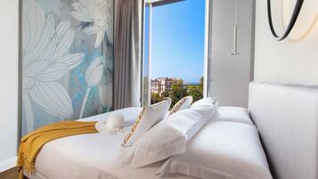 Habitación de hotel con una cama grande, ropa de cama blanca, vista al mar y un mural en la pared.