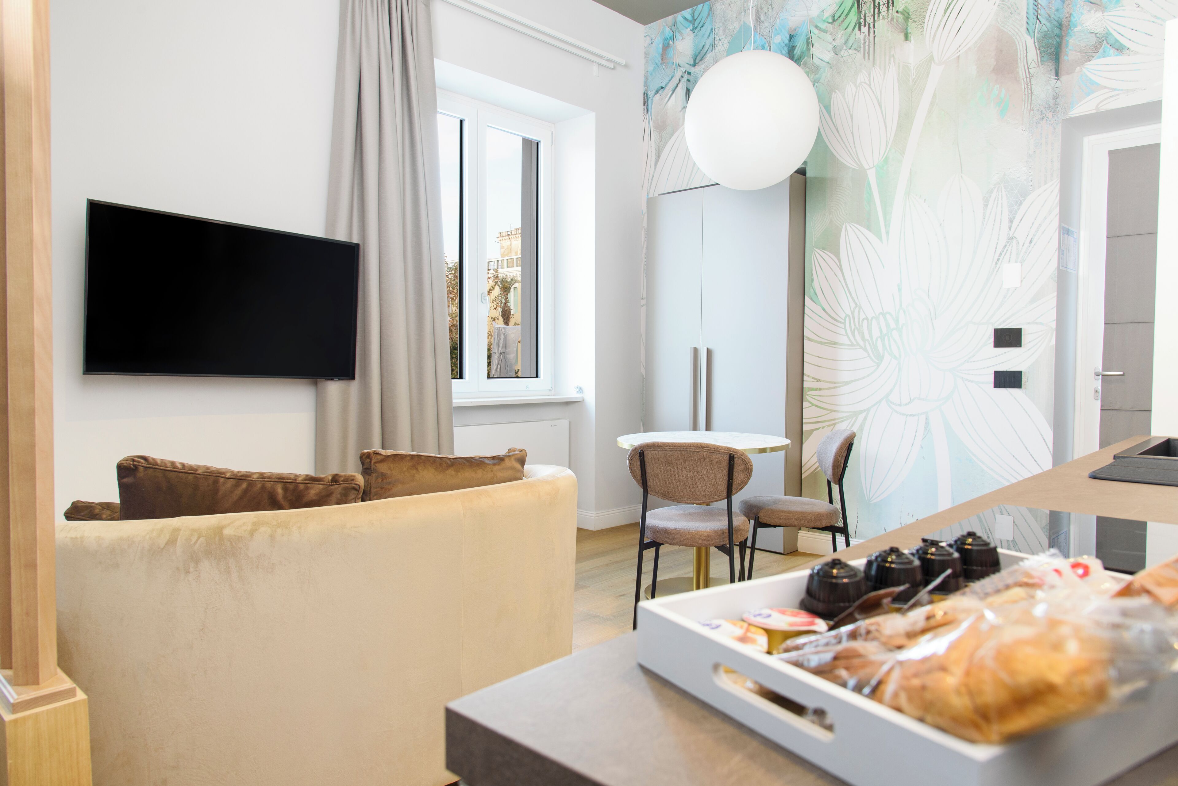 A modern living room with a flat-screen TV, a sofa, a dining table, and a wall with a floral mural.