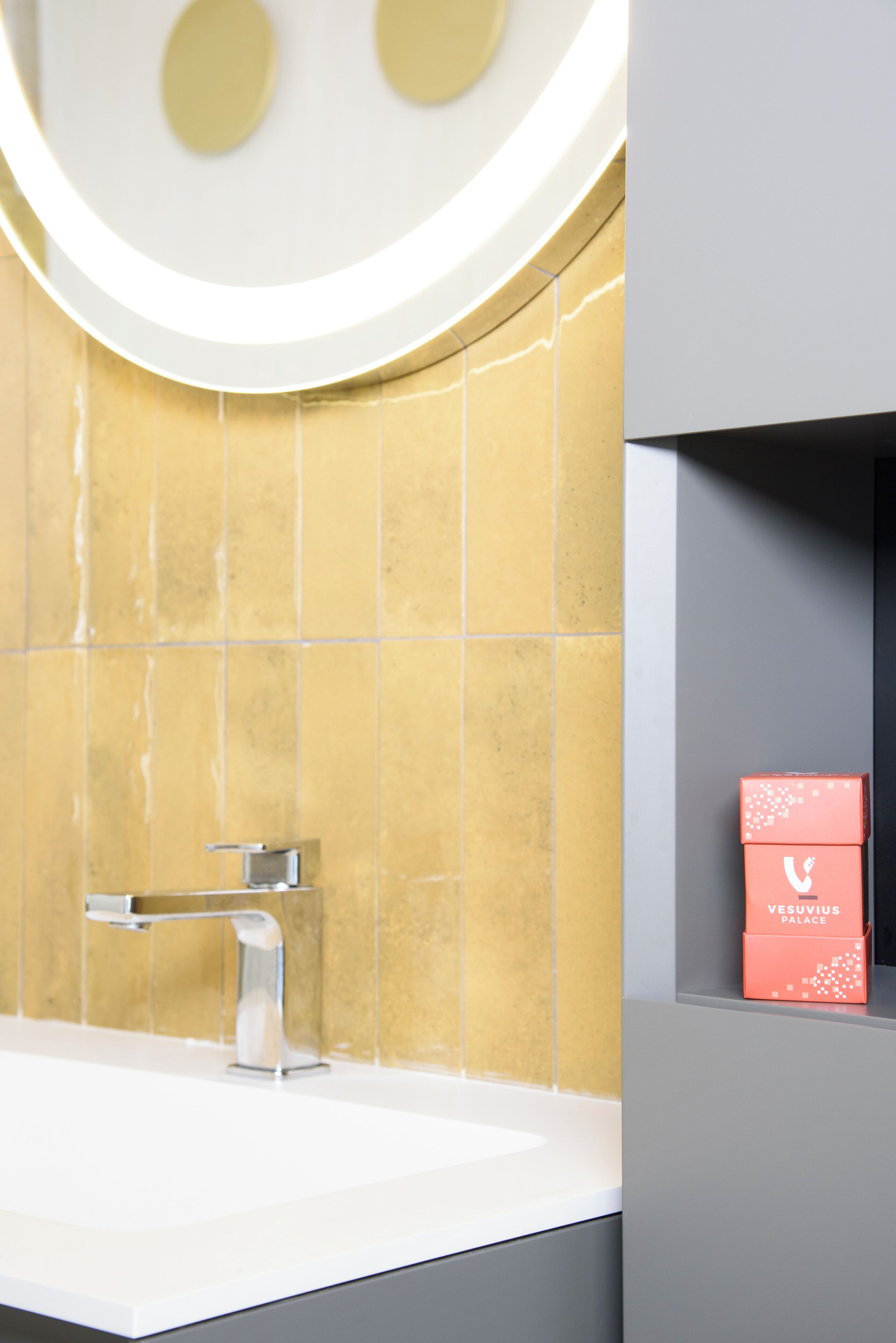 A bathroom with a white sink, a modern faucet, and a yellow-tiled wall. There is a red box labeled "VESUVIUS PALACE" on a shelf.