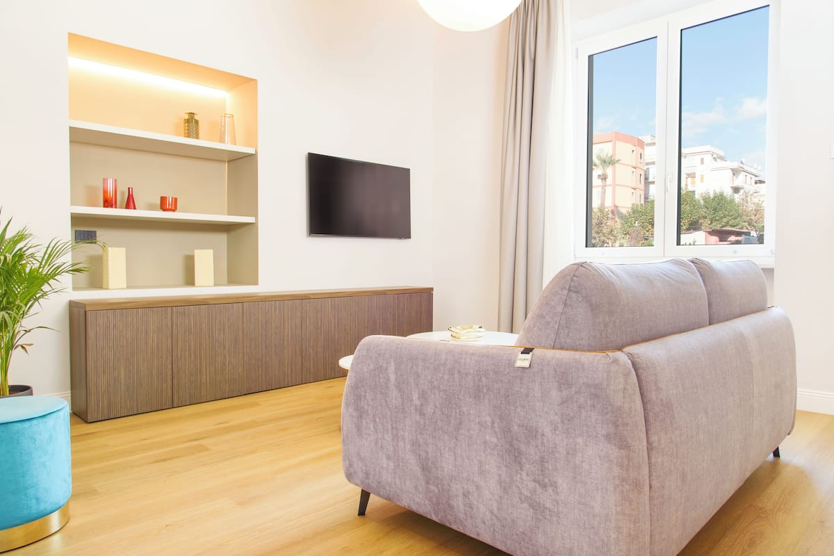 A modern living room with a sofa, a TV mounted on the wall, a wooden cabinet, and a window with a view of buildings.
