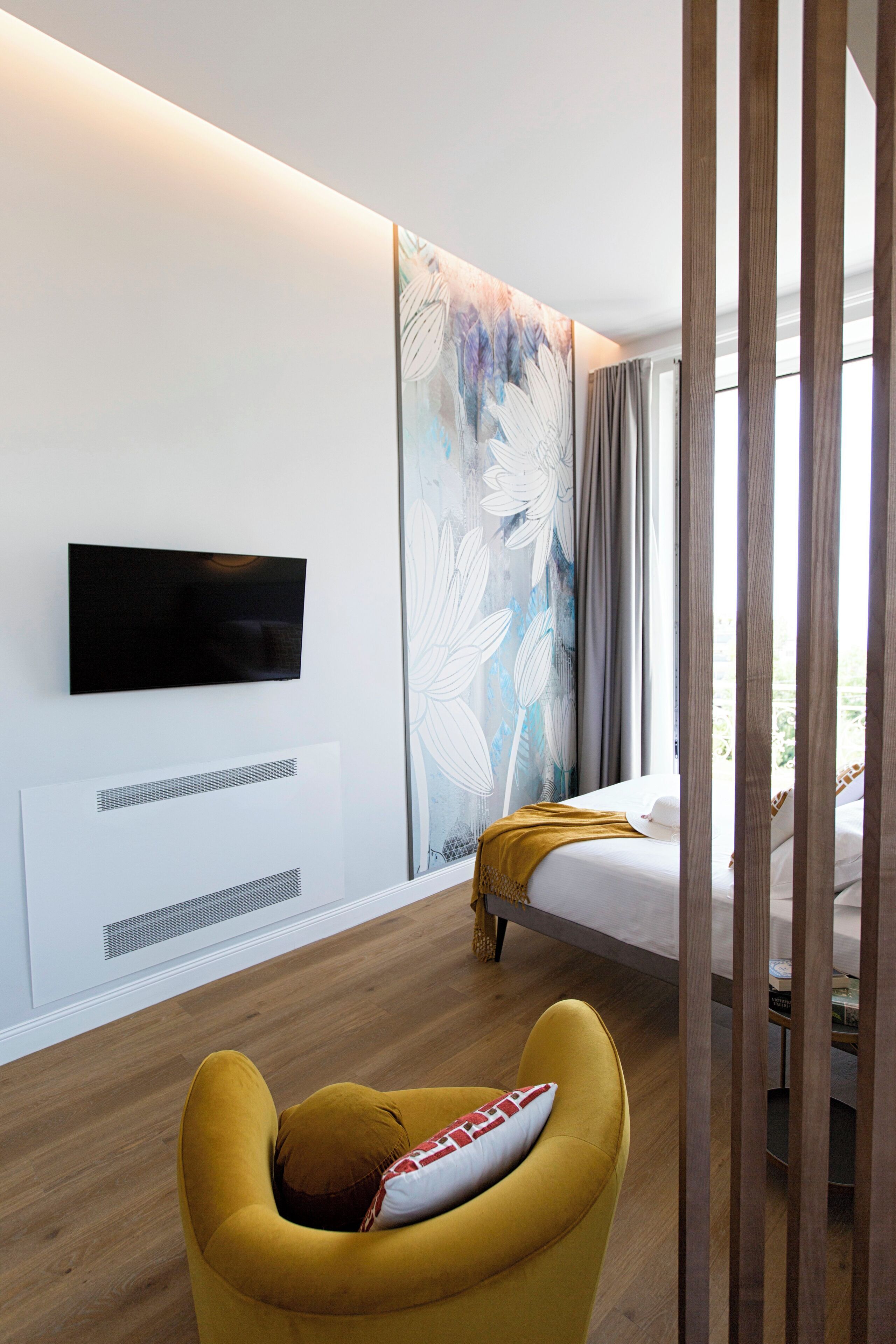 A modern hotel room with a flat-screen TV mounted on the wall, a bed with a mustard-colored throw, and a large floral artwork on the wall.