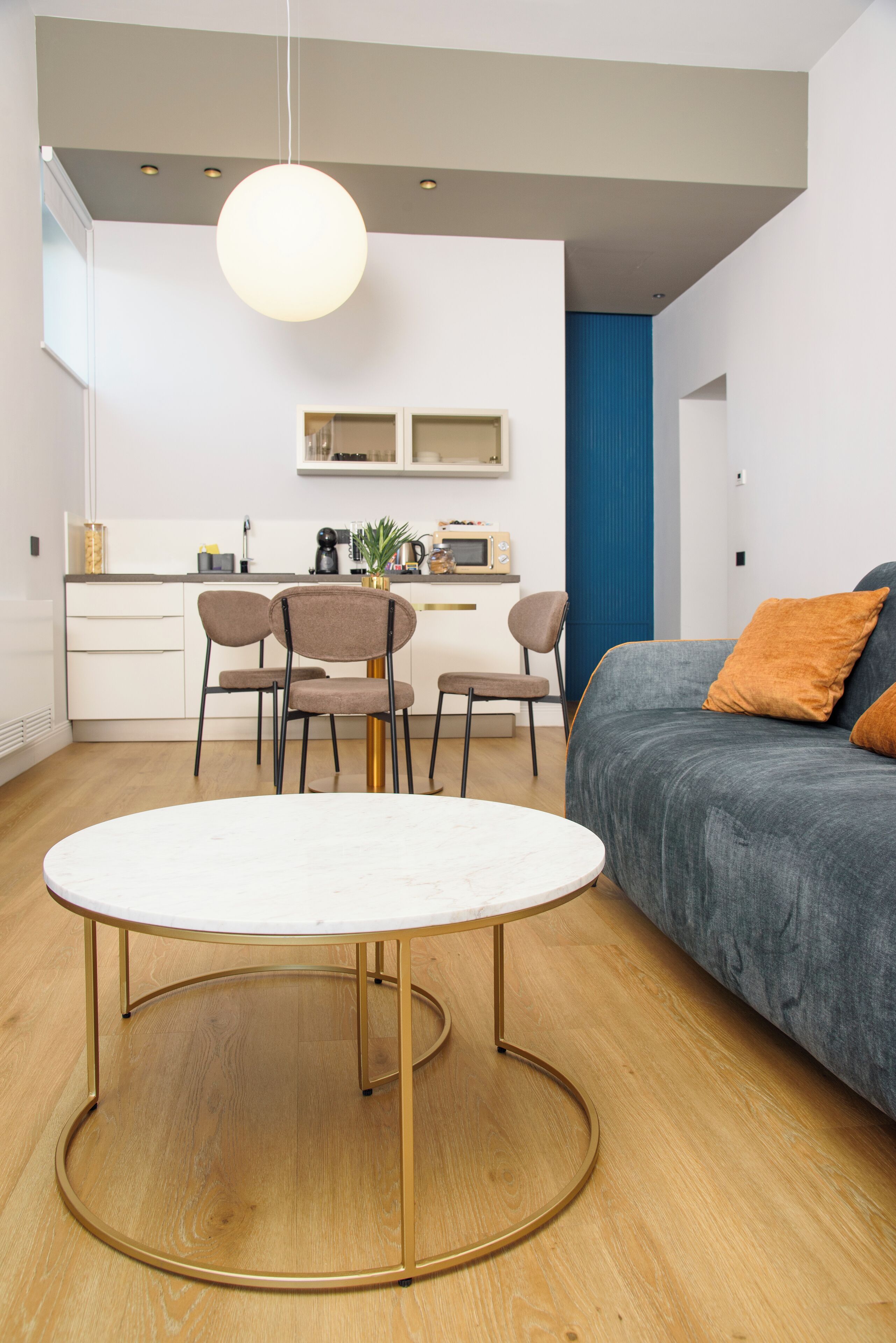 A modern living room with a round white table, two brown chairs, a grey sofa with orange cushions, and a blue accent wall.