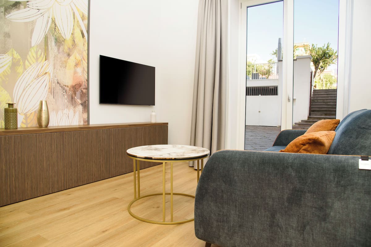 A modern living room with a flat-screen TV, a round coffee table, a grey sofa, and a view of the outside through a glass door.