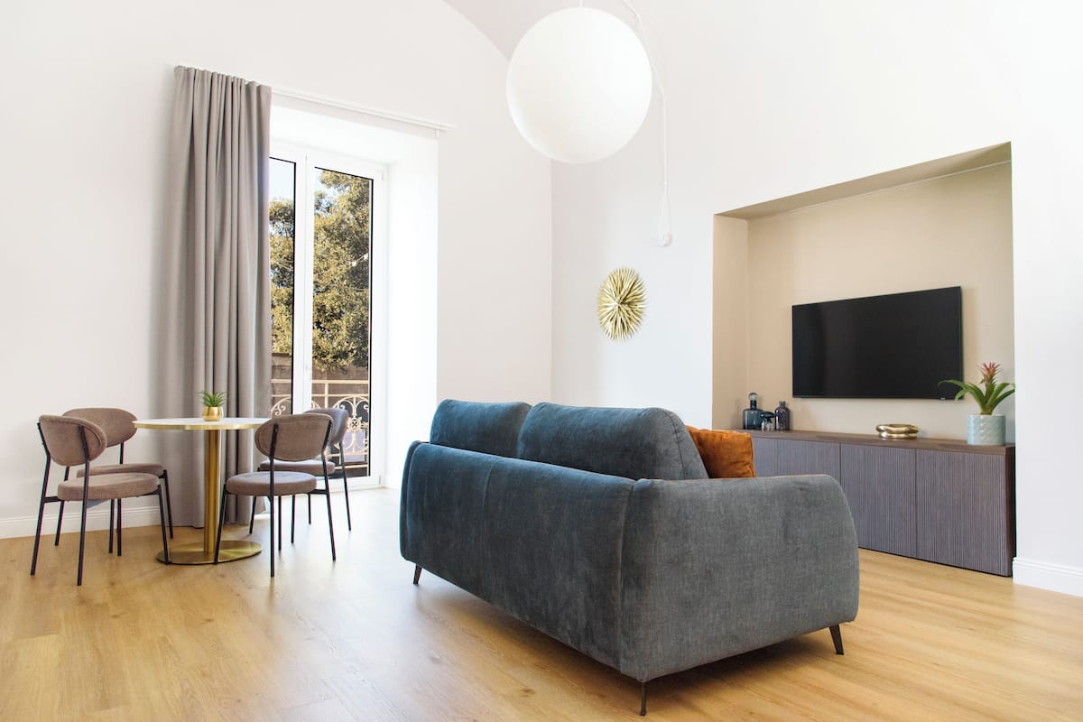 A modern living room with a grey sofa, a round table with chairs, a large window with curtains, a flat-screen TV, and a decorative wall piece.