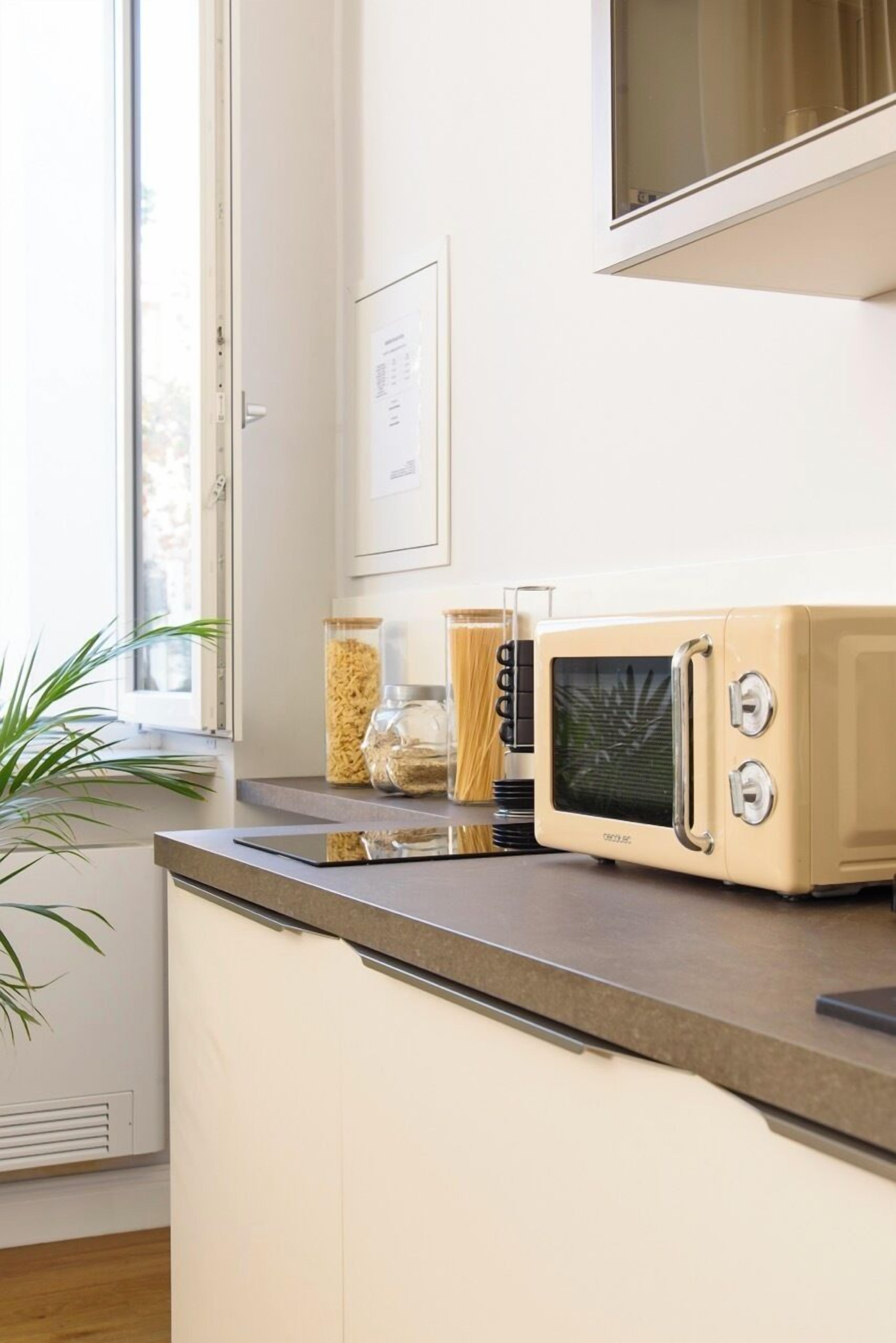 Una cocina con microondas, frascos de granos y una ventana con una planta.