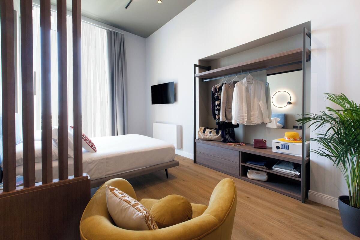 A modern hotel room with a bed, a yellow armchair, a wardrobe, a television, and a potted plant.
