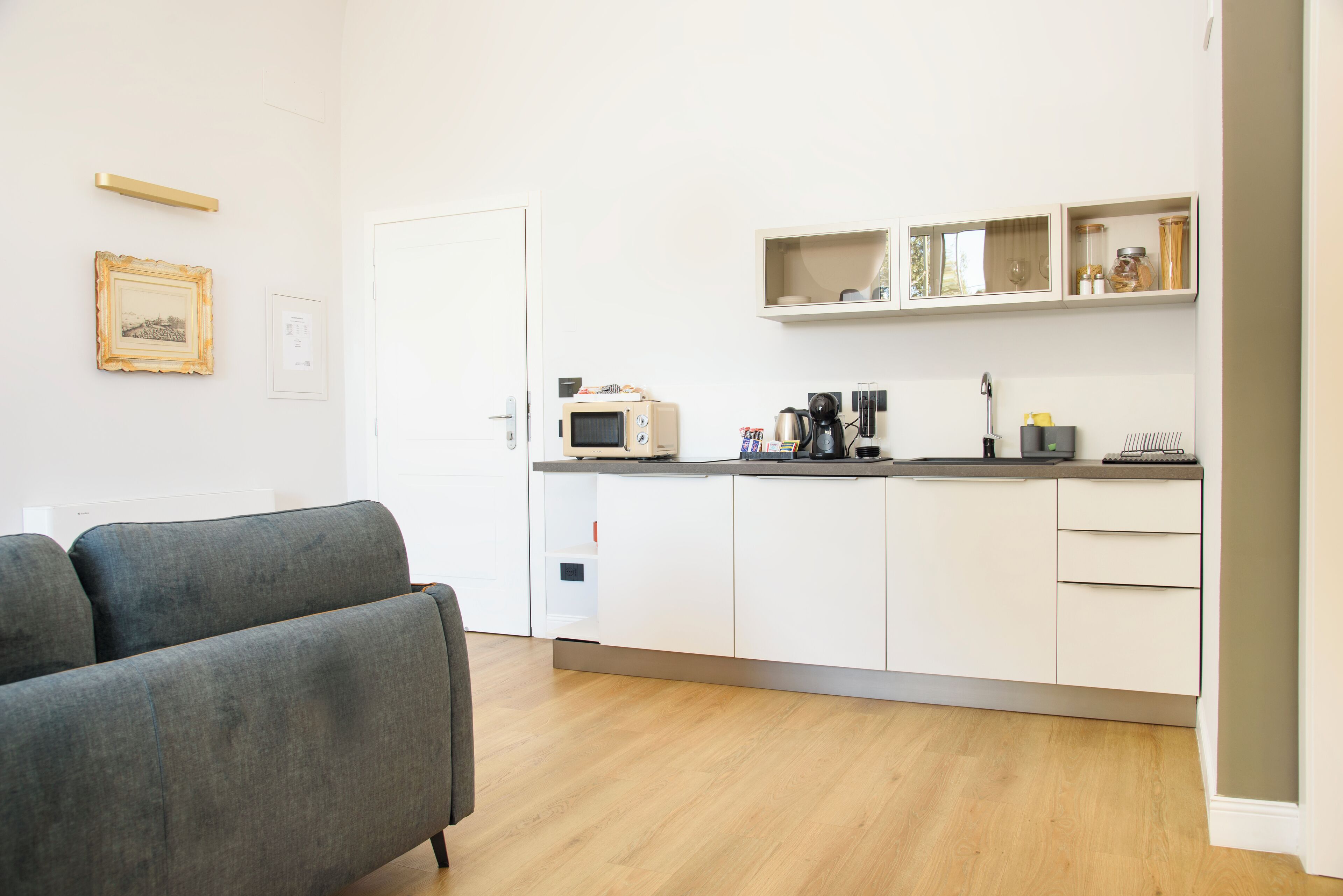 Een moderne keuken met witte kasten, een magnetron en een koffiezetapparaat. In de woonkamer staat een grijze bank.