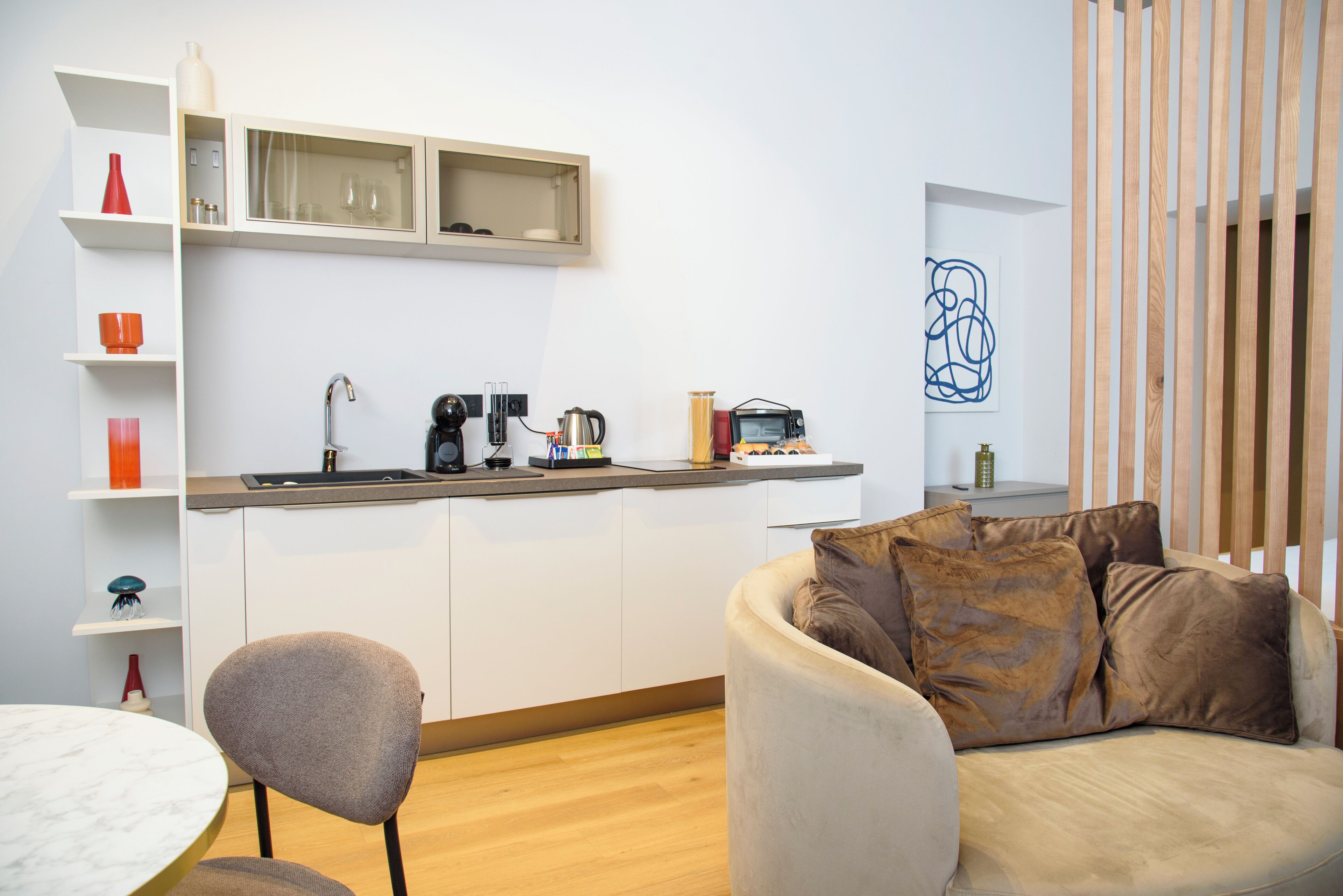 A modern kitchen with white cabinets, a coffee machine, and a small countertop. There is a dining area with a round table and chairs, and a sofa with brown cushions.