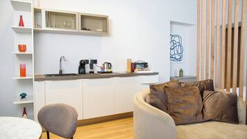 A modern kitchen with white cabinets, a coffee machine, and a small countertop. There is a dining area with a round table and chairs, and a sofa with brown cushions.