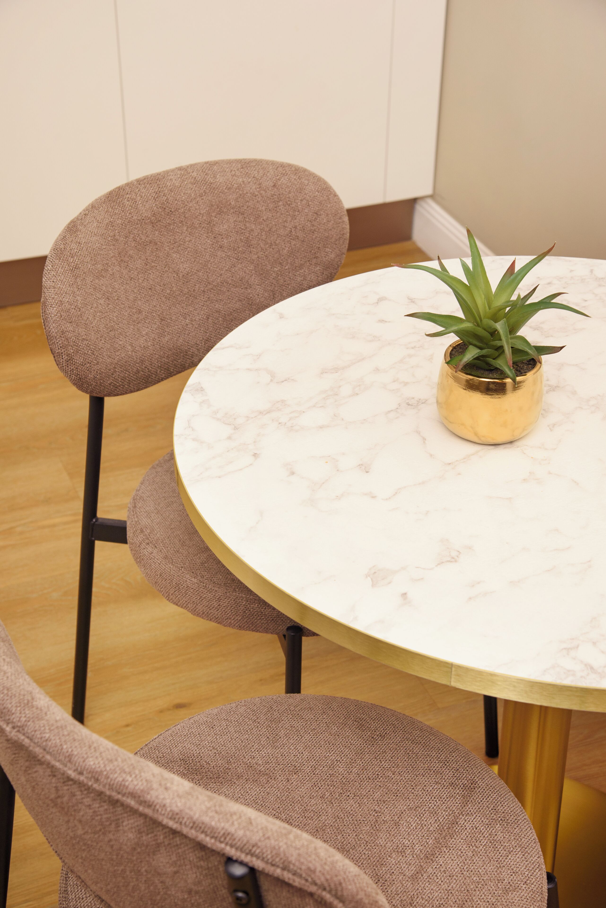 Una mesa redonda con tapa de mármol, rodeada por cuatro sillas tapizadas en marrón. Una planta en maceta está sobre la mesa.