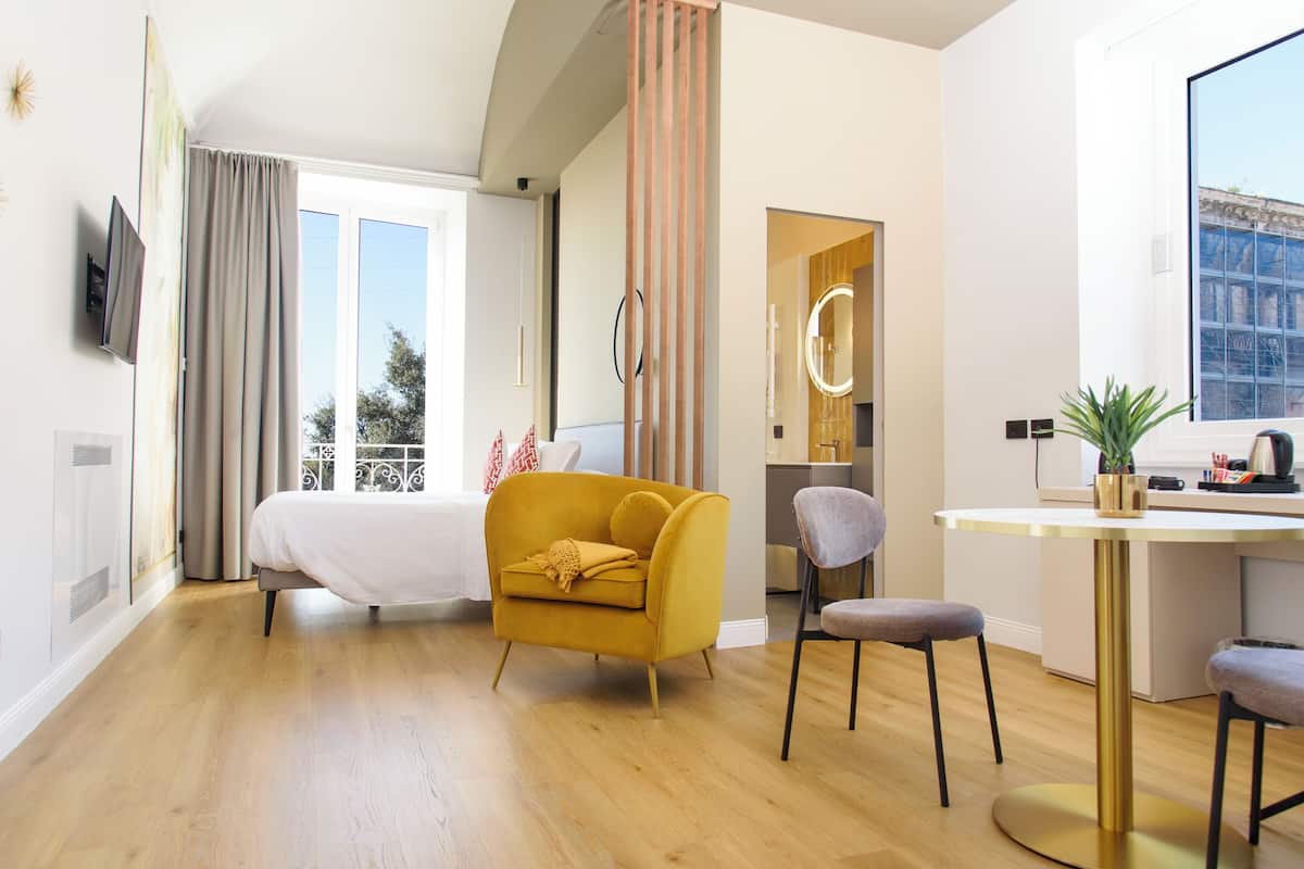 A modern hotel room with a bed, a yellow armchair, a desk, and a large window.