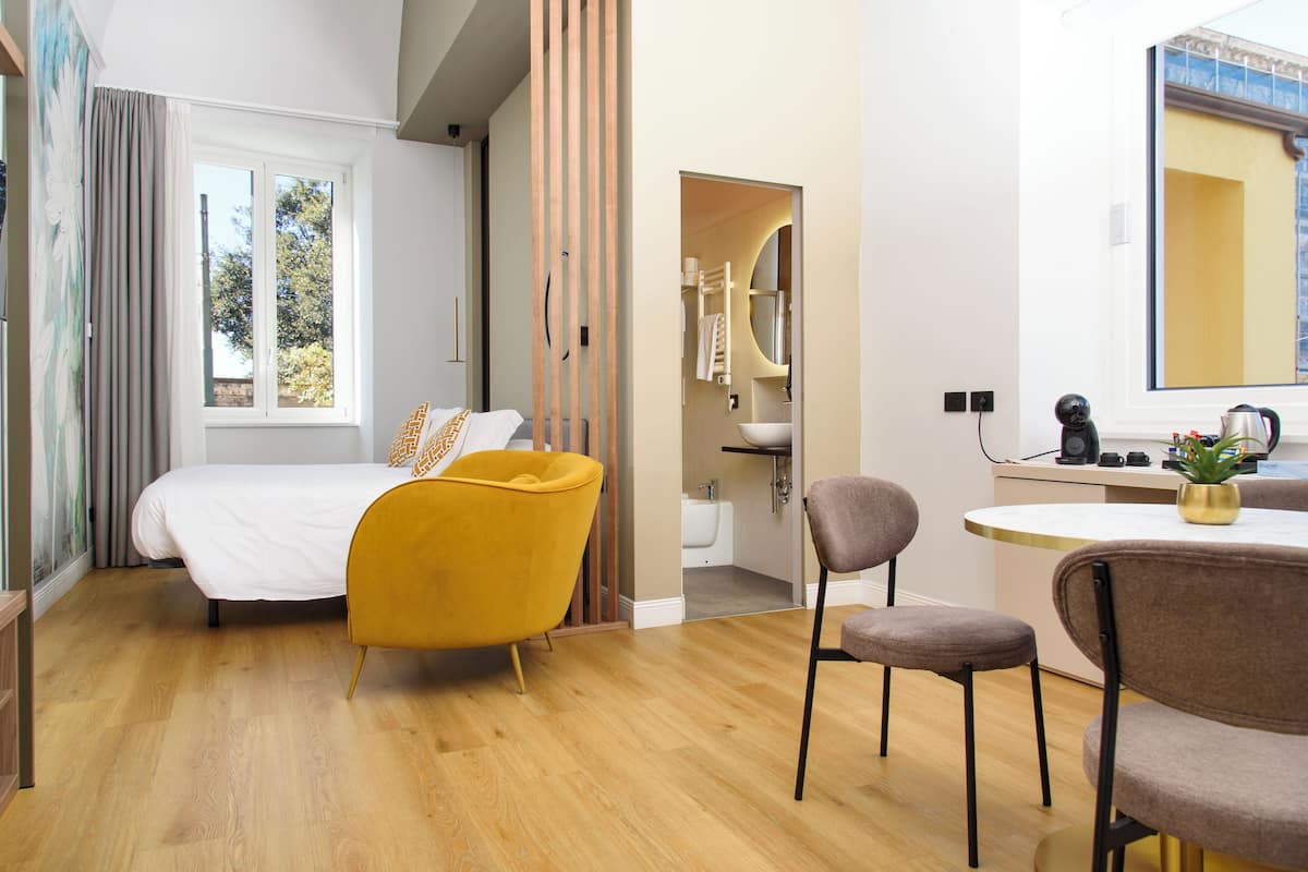 A modern hotel room with a bed, a yellow armchair, a gray chair, a small round table, and a bathroom visible through an open door.