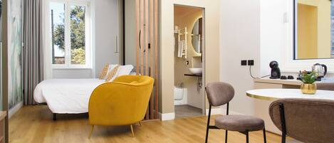 A modern hotel room with a bed, a yellow armchair, a gray chair, a small round table, and a bathroom visible through an open door.
