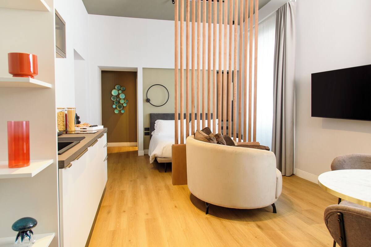 A modern hotel room with a wooden floor, a round dining table, a sofa, a bed with a headboard, and a TV mounted on the wall.