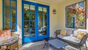 A sunlit patio with a blue door, wicker furniture, a patterned rug, and a decorative window grille.