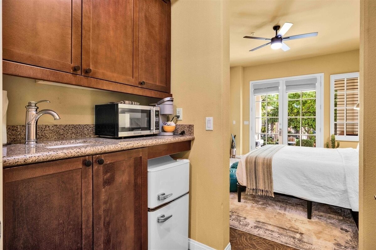 A compact kitchen with wooden cabinets, a microwave, and a small refrigerator. Adjacent to the kitchen is a bedroom with a bed, window, and a ceiling fan.