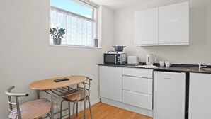 Eine kompakte Küche mit Mikrowelle, Toaster, Wasserkocher und einem kleinen Kühlschrank. Ein Fenster mit einem Blumentopf auf der Fensterbank, und ein Hocker an einem Tisch.