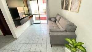 A living room with a grey sofa, a flat-screen TV mounted on the wall, a sliding glass door leading to an outdoor area, and a potted plant. - Feet in the Sand Corner of the Fort III (Praia Grande)