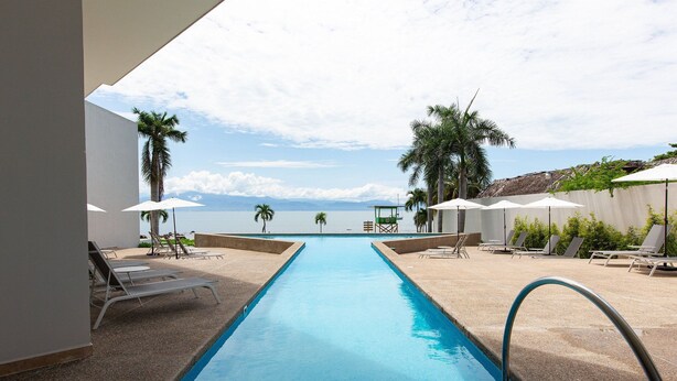 A swimming pool with lounge chairs and umbrellas, surrounded by palm trees and a view of the ocean. - Beautiful beachfront Studio, Zantamar 304 (Cruz de Huanacaxtle)