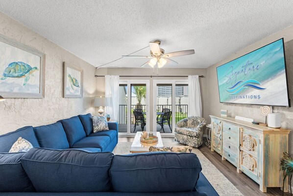 A living room with a blue sofa, a ceiling fan, a TV on the wall, and a view of the outdoors through glass doors.