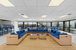 Un espace intérieur vaste et lumineux, comprenant un grand canapé d’angle, deux tables basses et de grandes fenêtres offrant une vue dégagée.