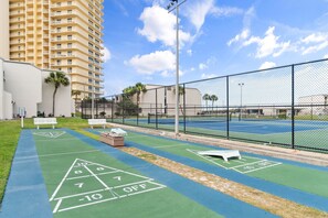 Una cancha de tenis con una cerca de malla metálica, una cancha de juego de bolos verde y un edificio alto al fondo.