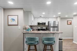 Eine moderne Küche mit weißen Schränken, einem Edelstahl-Kühlschrank, einer Mikrowelle und einer Arbeitsplatte aus Granit. Zwei türkisblaue Barhocker und ein Mülleimer sind ebenfalls vorhanden.