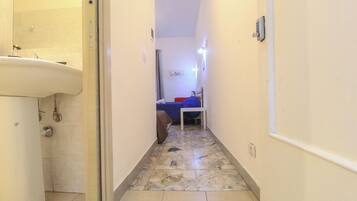 A narrow hallway with a bathroom on the left, a bed with a blue cover in the center, and a white door on the right.
