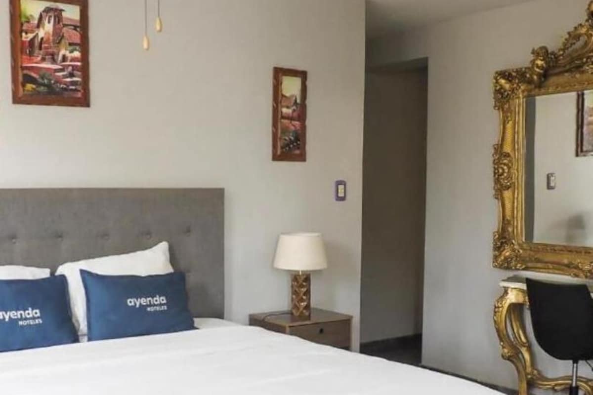 Una habitación de hotel con una cama grande, un escritorio, una silla, un ventilador de techo y dos cuadros en la pared.