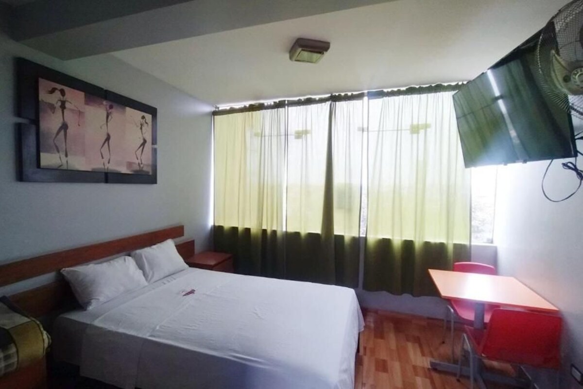 Una habitación de hotel con cama, escritorio, silla, ventana con cortinas y ventilador de techo.