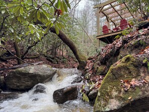 A wooden structure with red Adirondack chairs overlooking a stream in a forested area.